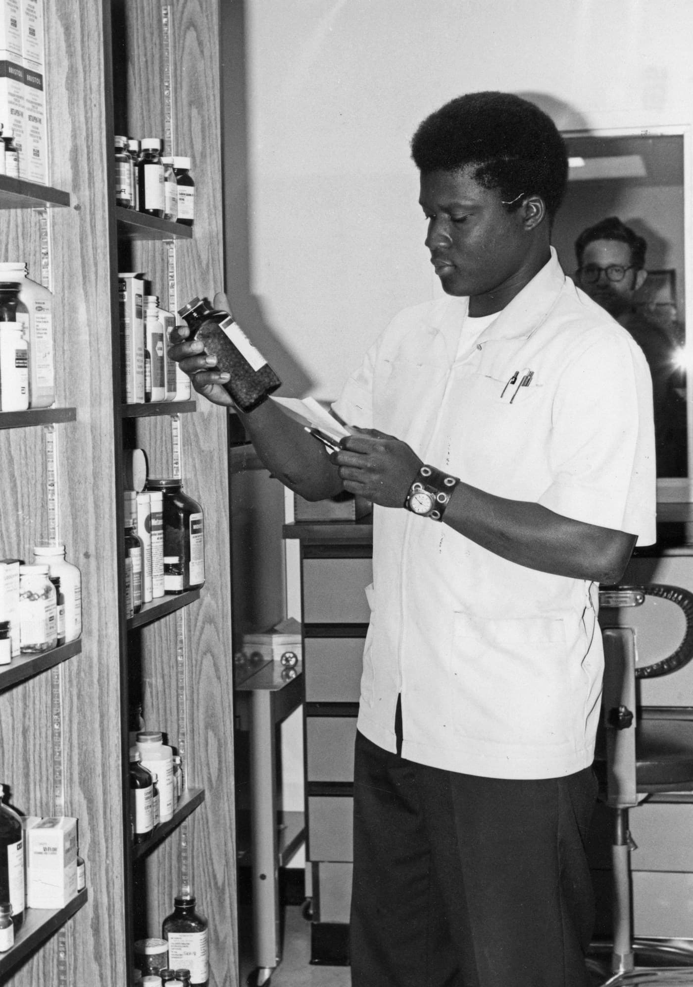 A pharmacist checking a prescription against a stock bottle, photographed in black and white